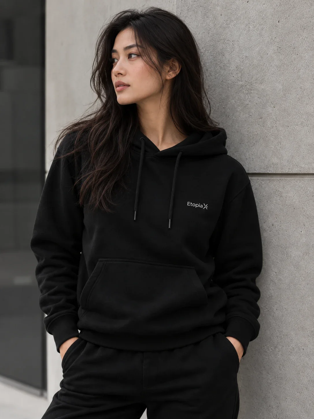 Le sweat à capuche noir EtopiaX porté par une femme, adossée à un mur de béton